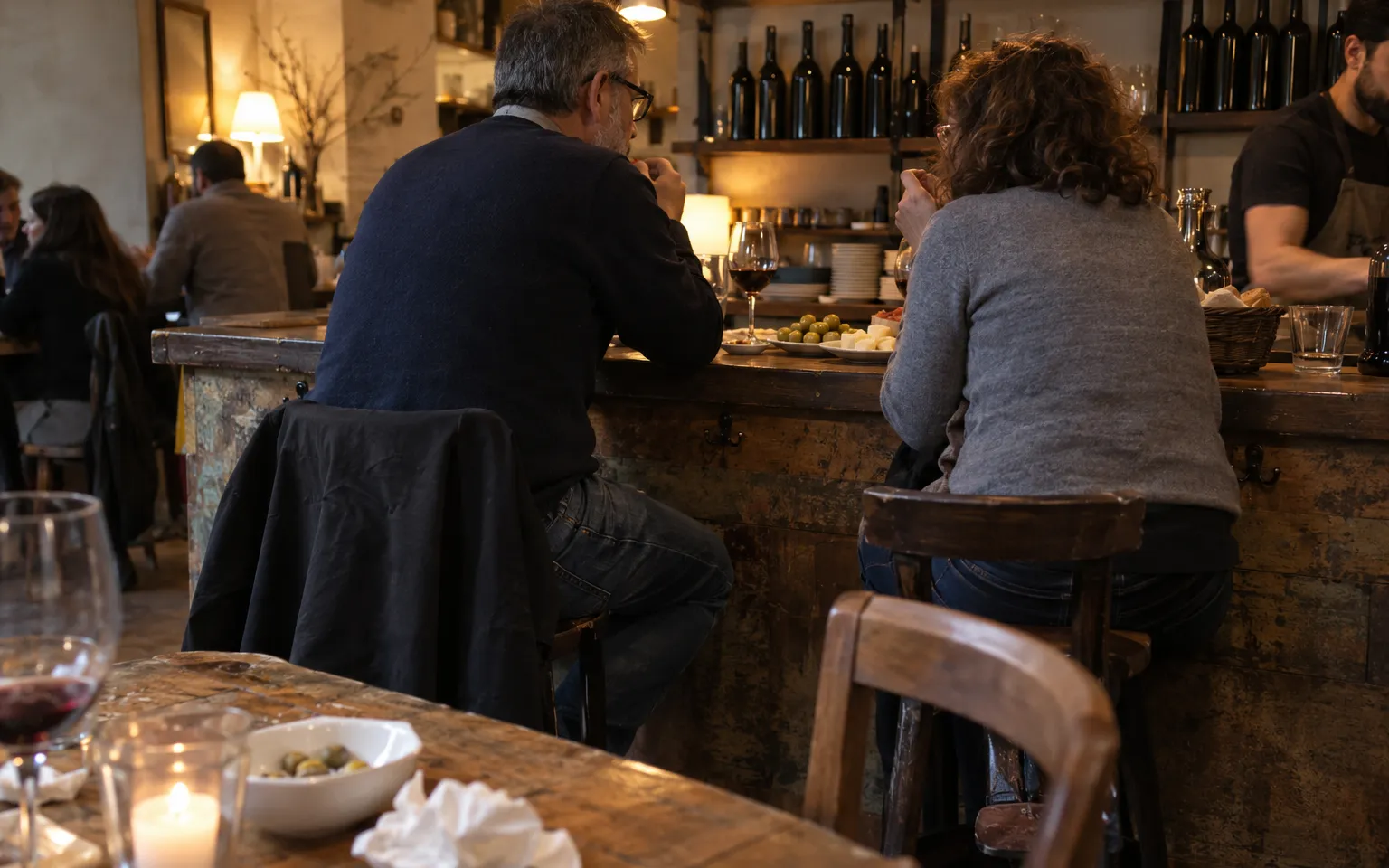 Piccolo bistrot con bancone, bottiglie di vino e due clienti agli sgabelli